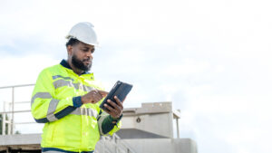 A man in a yellow safety jacket is looking at a tablet. He is wearing a hard hat and safety glasses. electrical, engineer, environment, energy, clean, sustainable, wind turbine, nature