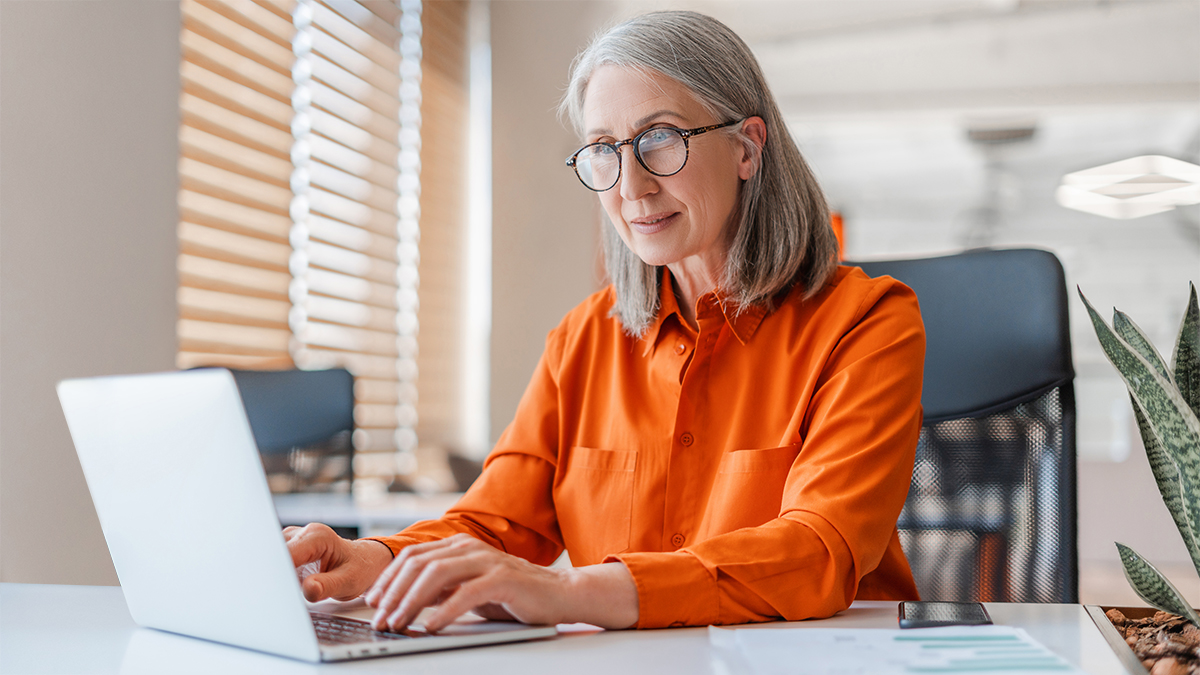 Smiling mature businesswoman wearing stylish eyeglasses, orange shirt using laptop working online in modern office. Manager, writer planning project, typing on keyboard sitting at workplace