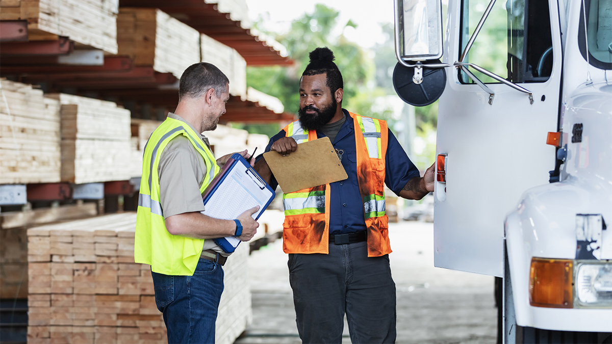 Two multiracial men in lumberyard, with clipboards, truck